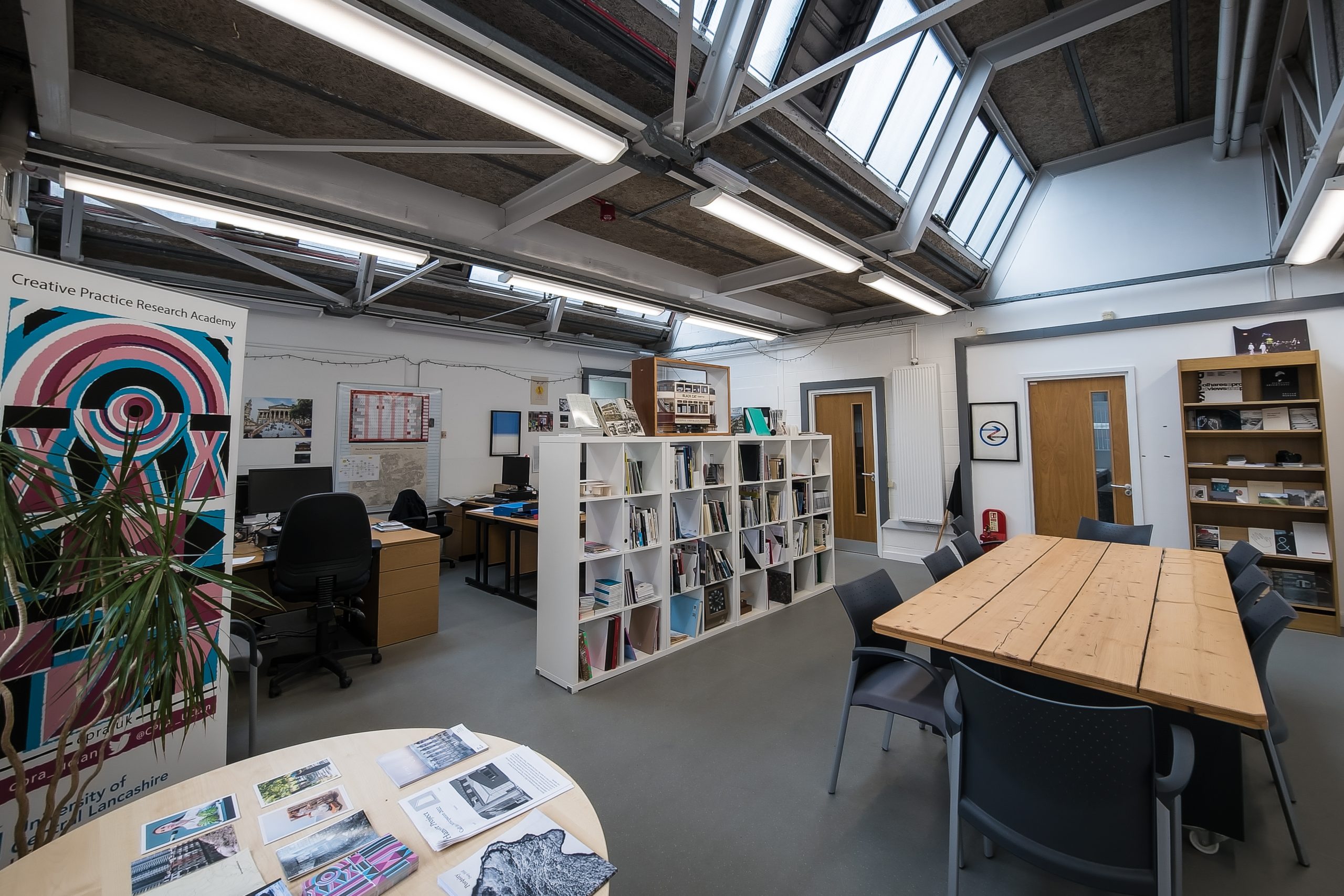 Photo of a large office, with stirp lighting and skylight windows. There is a colourful banner to the left of the image and a long table on the right, made from wooden planks. The room is divided by a large bookcase, filled with books and with a model tram in a Perspex case on the top.