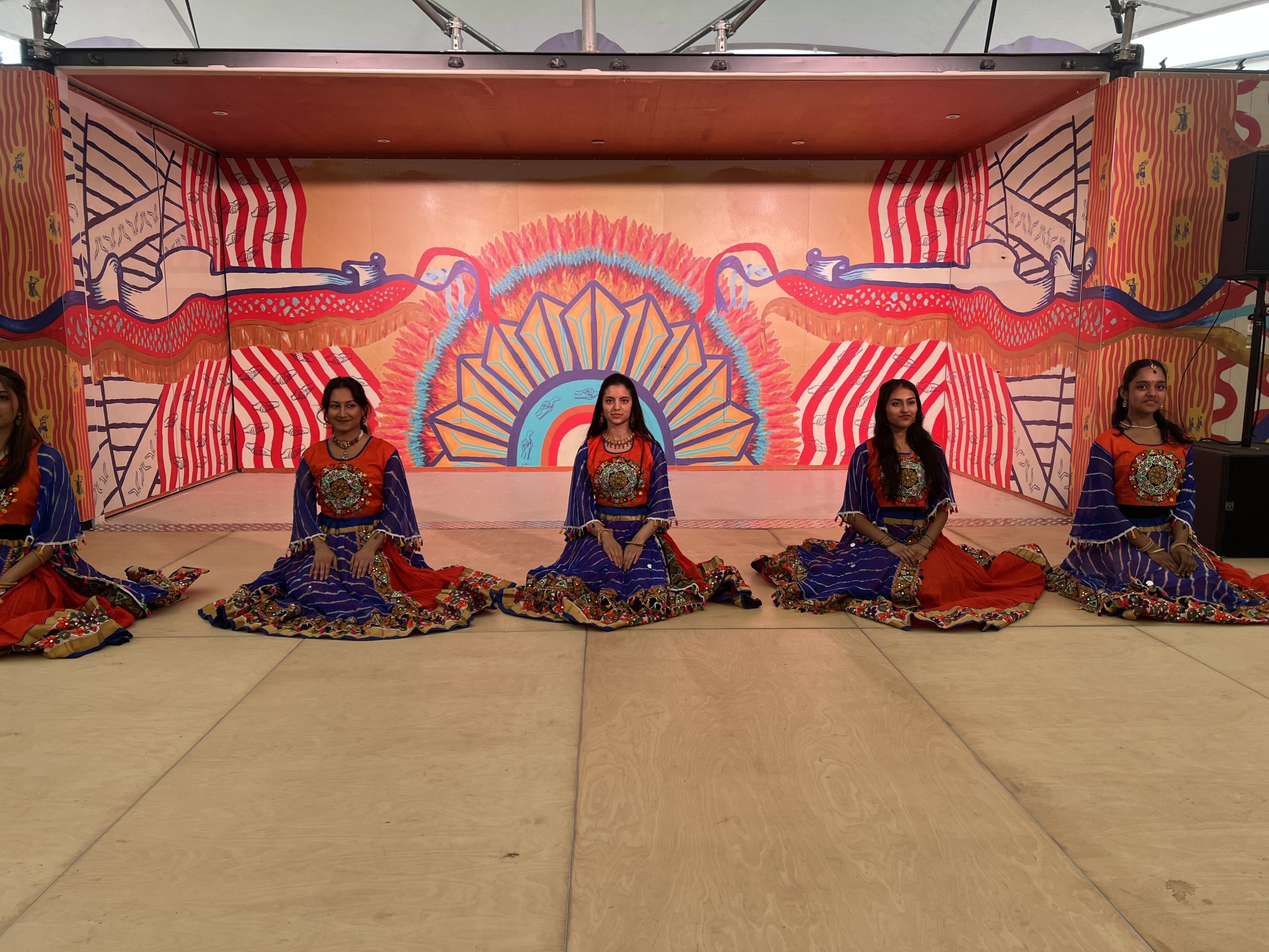 Photograph of five women of South Asian heritage, seated in a line in front of a brightly coloured background. The women are all wearing matching red and purple dresses, which are embroidered with gold thread and colourful beads.