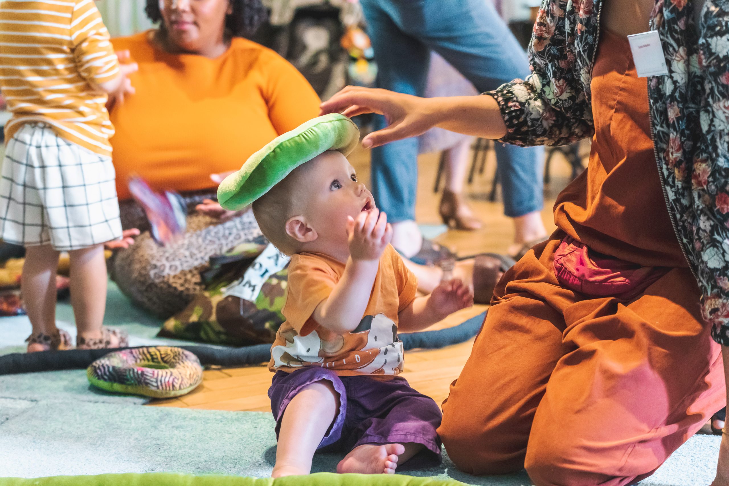 Photo of a toddler seated on a rug with a green doughnut shaped cushion on his head, looking up at a woman, whose face is out of the frame.