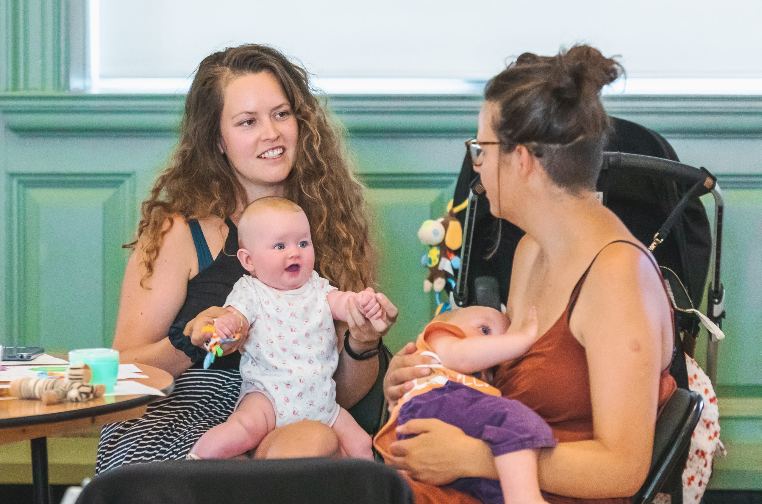 Photograph of two seated women. One has a baby on her lap and another is breastfeeding a baby.