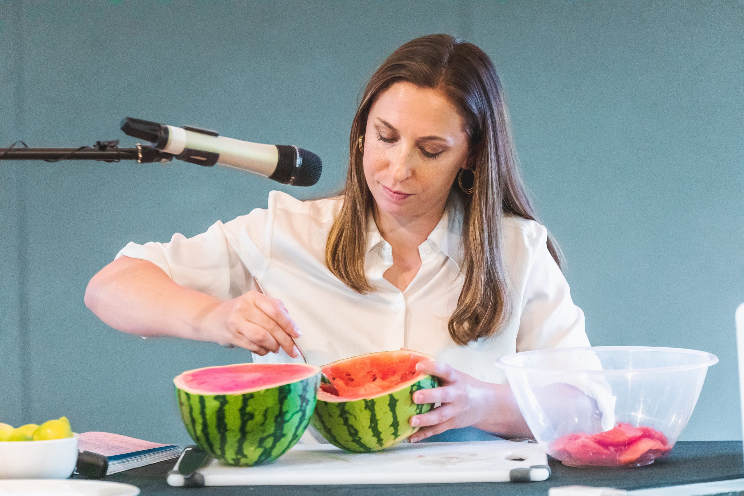 Photo of a woman seated at a table, in front of a microphone, scooping the flesh out of a watermelon, which has been cut in half, into a plastic bowl.
