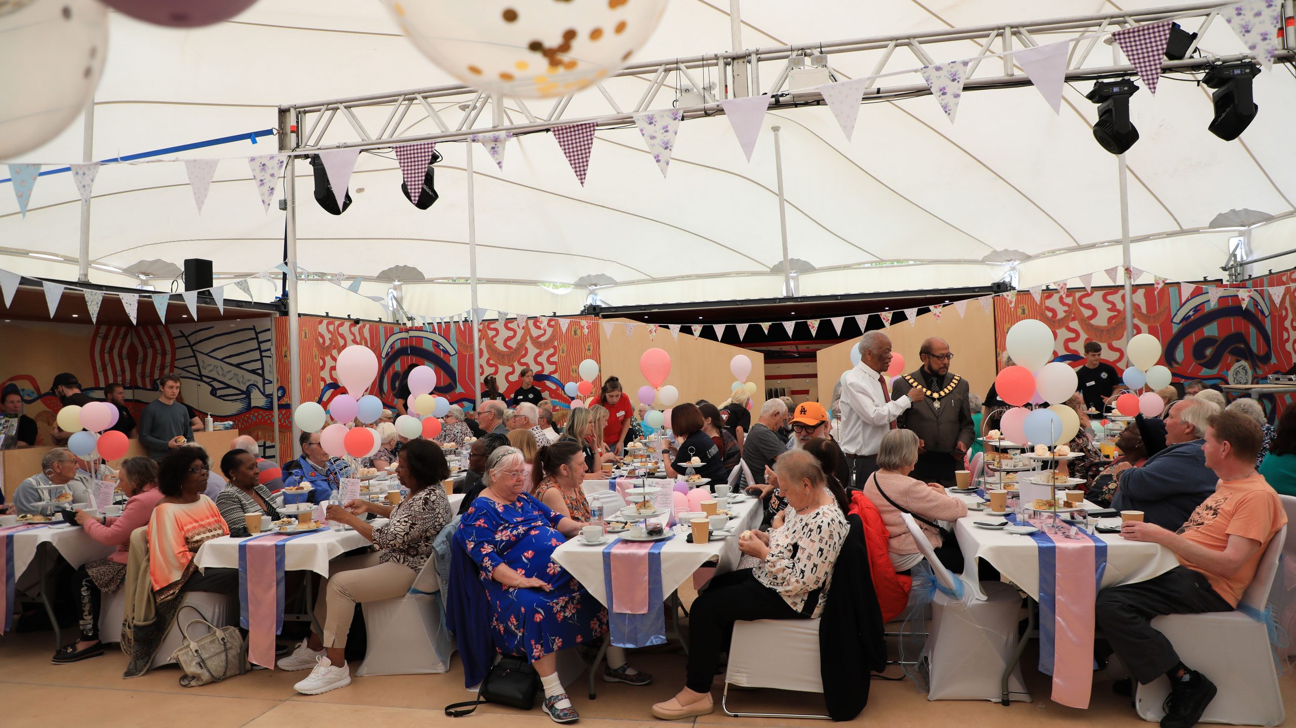 A photograph of the Afternoon Tea event taking place inside the MET. Attendees are sitting at 4 row of table enjoying cake and tea.