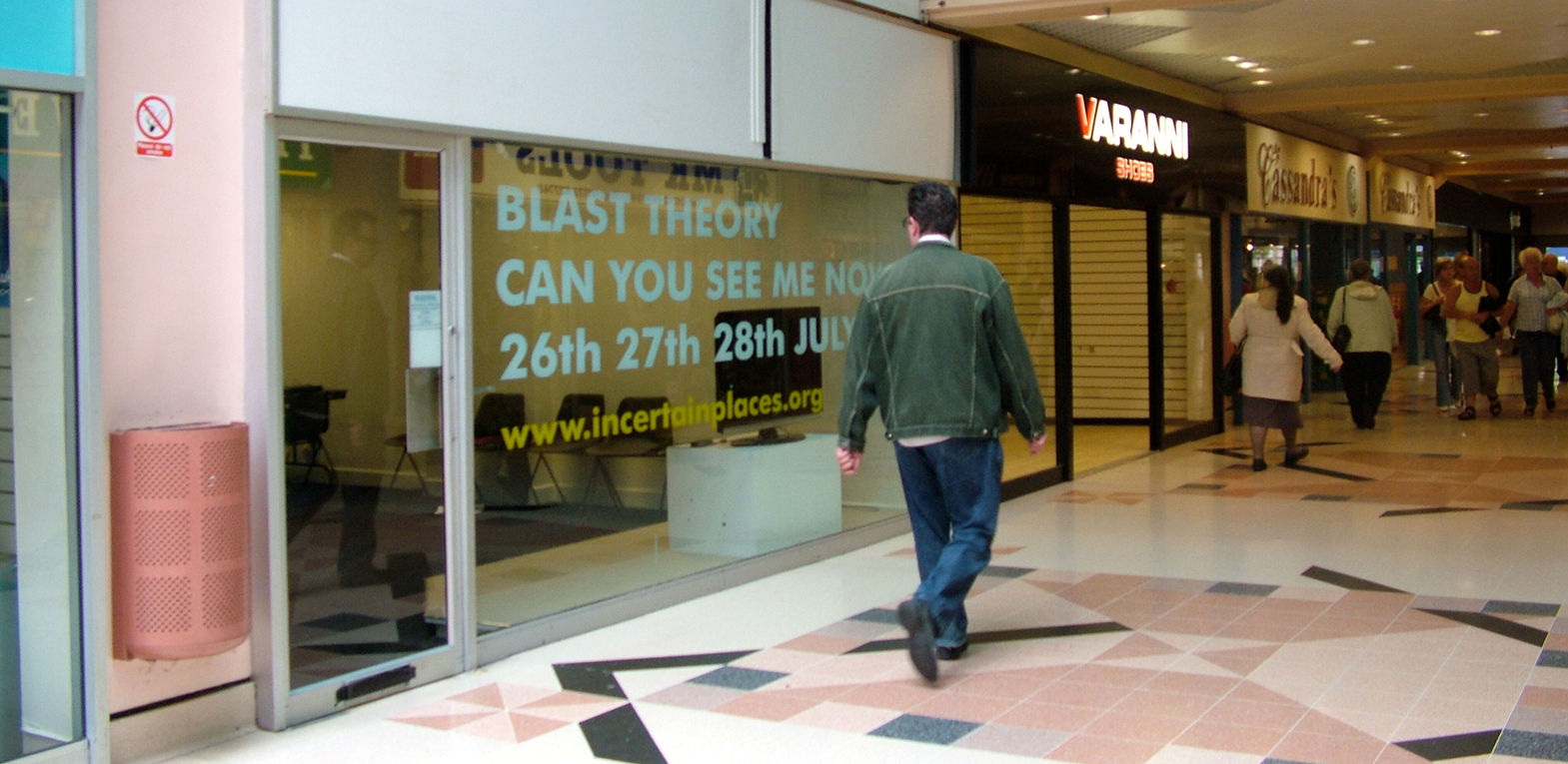 A man walks past a shop unit with the text displayed in the window 'Blast Theory, Can you see me now' and the dates.