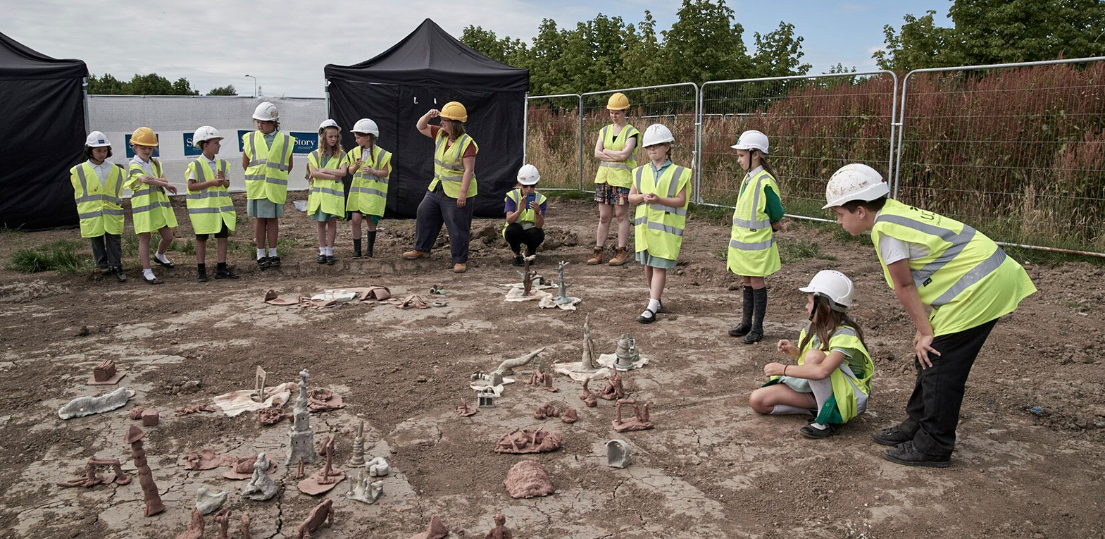 A group of children in hi-vis. led by the artist survey the model village that they have created out of clay