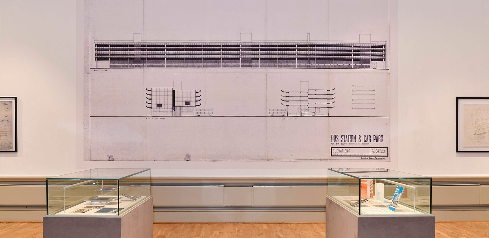 The exhibition in the Harris Museum. Artifacts from the bus station in display cases including models, pamphlets and the digital portion of the original clock. They are in front of a wall display of plans and architectural drawings.