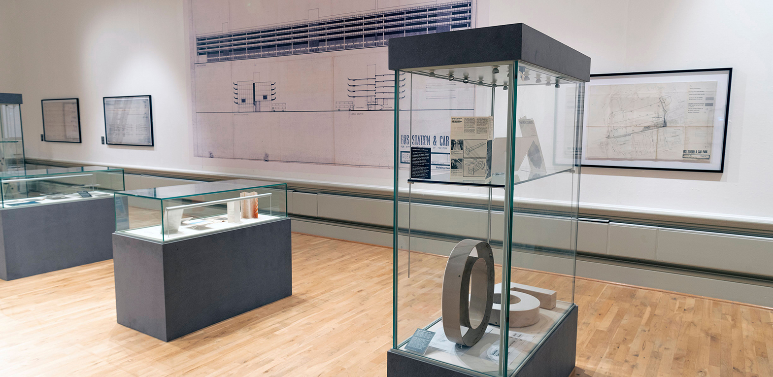 The exhibition in the Harris Museum. Artifacts from the bus station in display cases including models, pamphlets and the digital portion of the original clock. They are in front of a wall display of plans and architectural drawings.