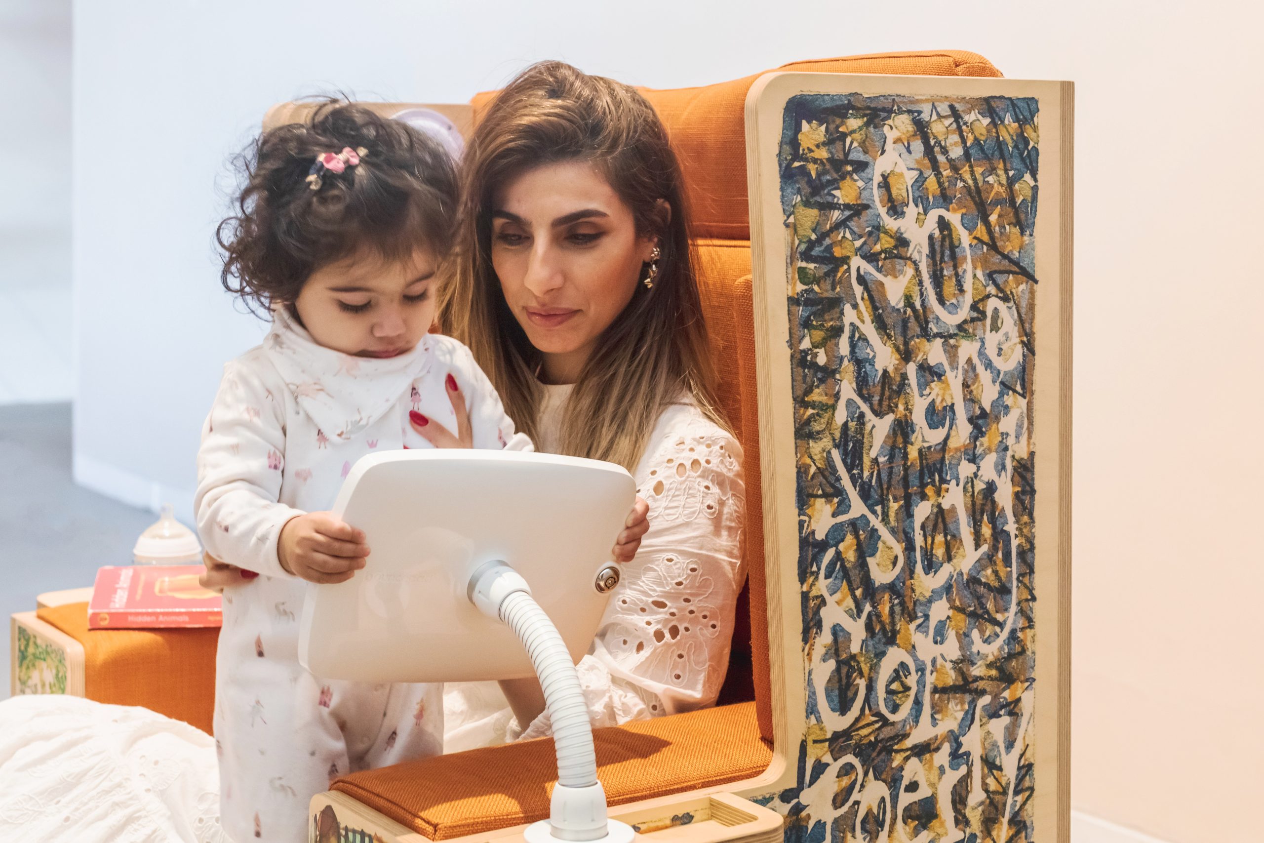 Photograph of a woman with long brown hair sitting in a large chair with a baby on her lap. The baby and woman are both dressed in white and looking at an iPad attached to the chair. The chair has a wooden frame and is decorated with drawings.