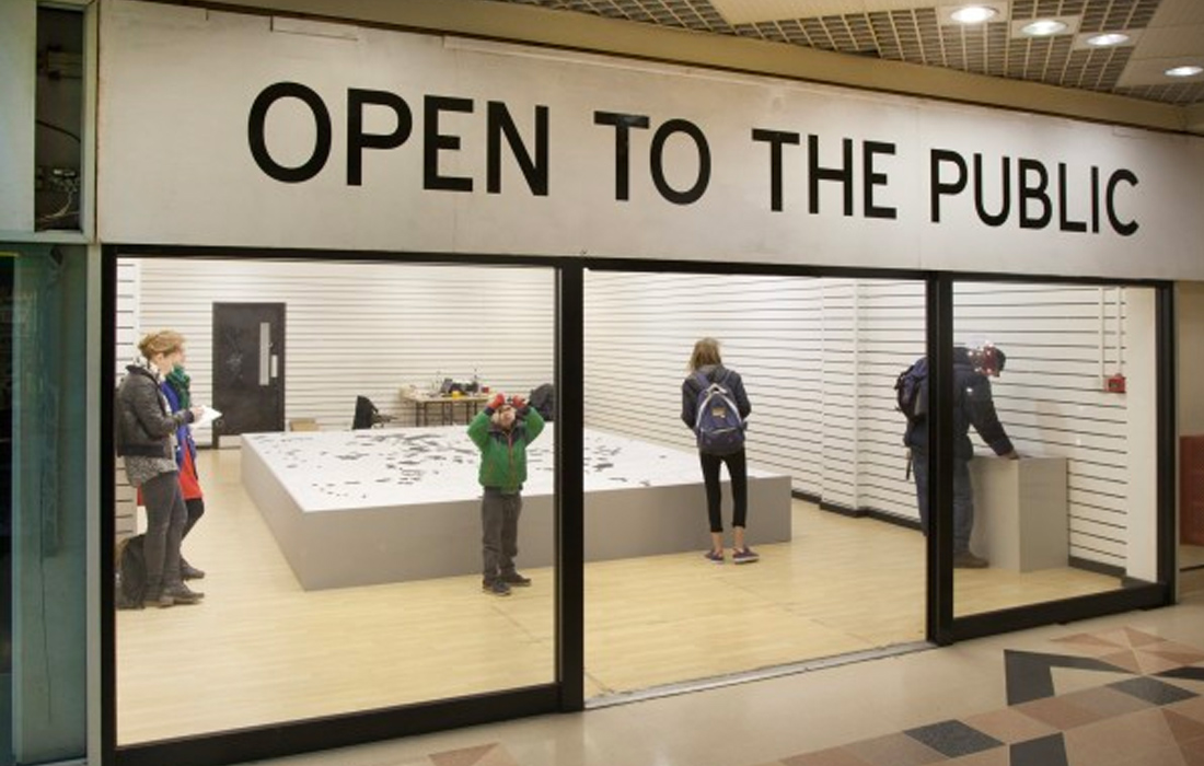 A empty shop exhibition for the 'Open to the Public' project. Visitors stand next to a plinth with a map of Preston city centre on it. On the map empty or disused buildings have been highlighted in black.
