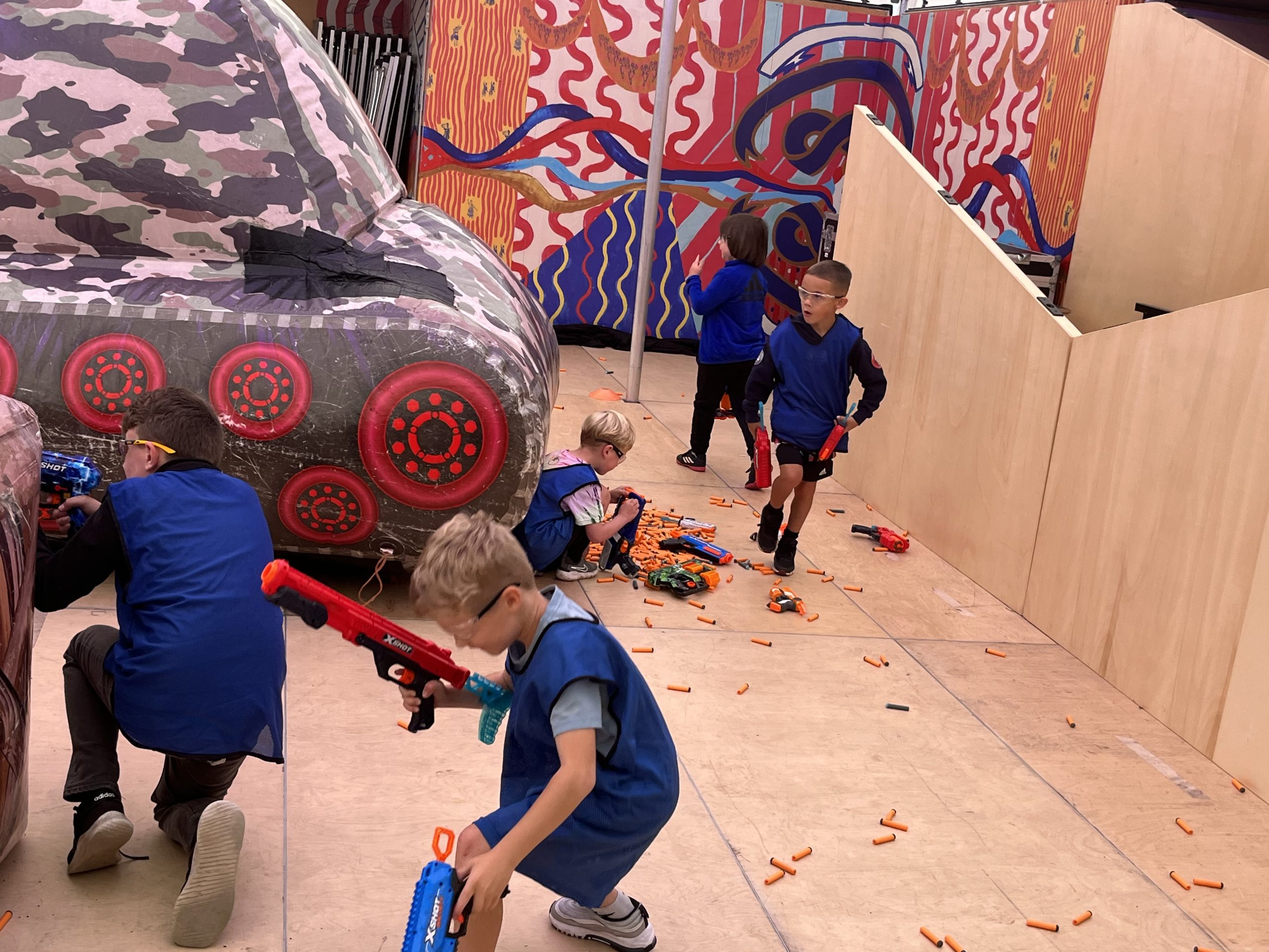 A photograph of a nerf party taking plac inside the MET. Several children playing with nerf guns and hiding behind inflatable obstacles.